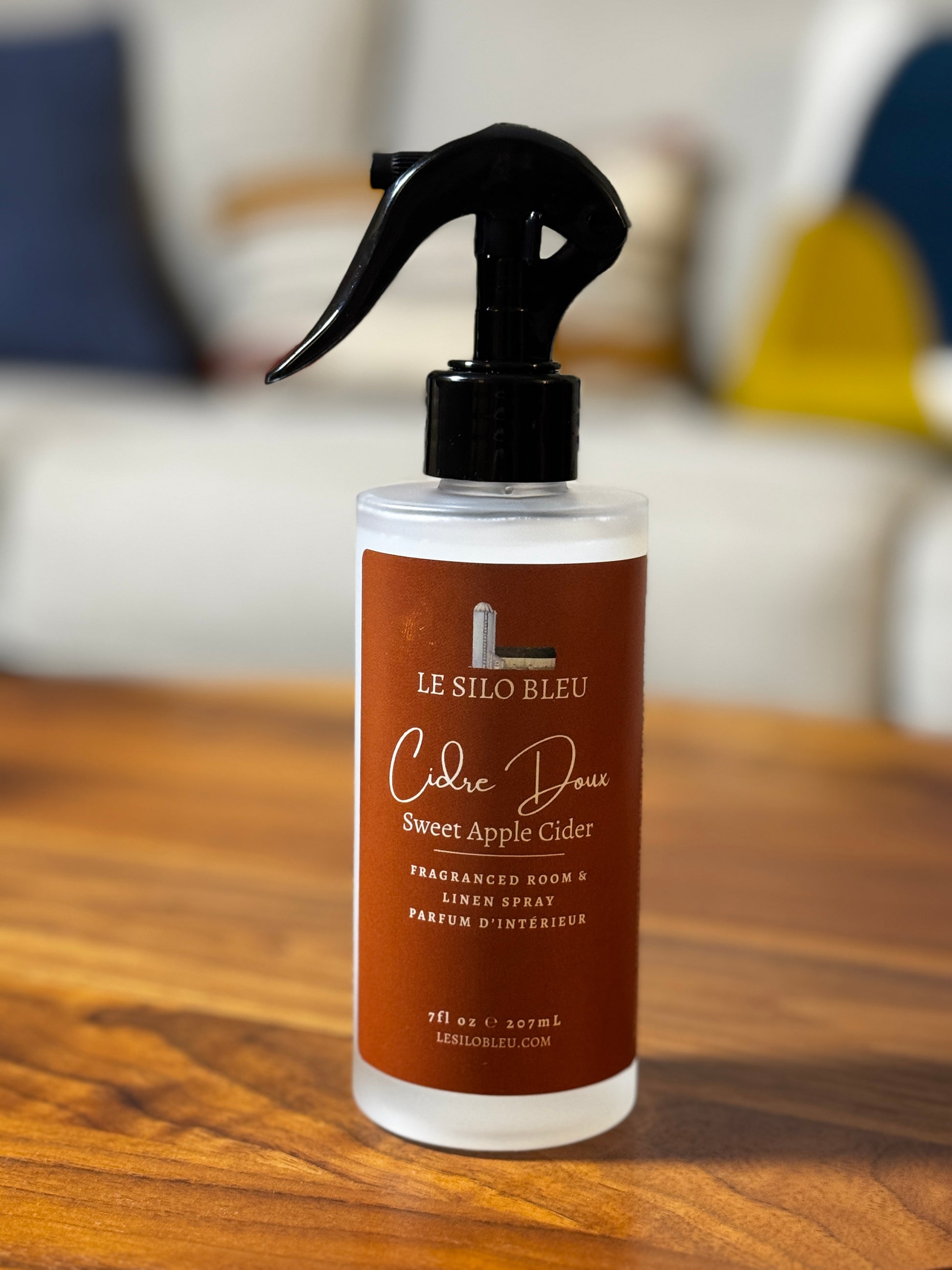 A bottle of Le Silo Bleu Sweet Apple Cidre room and linen spray, featuring a black trigger sprayer and a brown label, set on a wooden table with a blurred living room background.