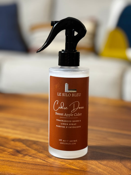 A bottle of Le Silo Bleu Sweet Apple Cidre room and linen spray, featuring a black trigger sprayer and a brown label, set on a wooden table with a blurred living room background.