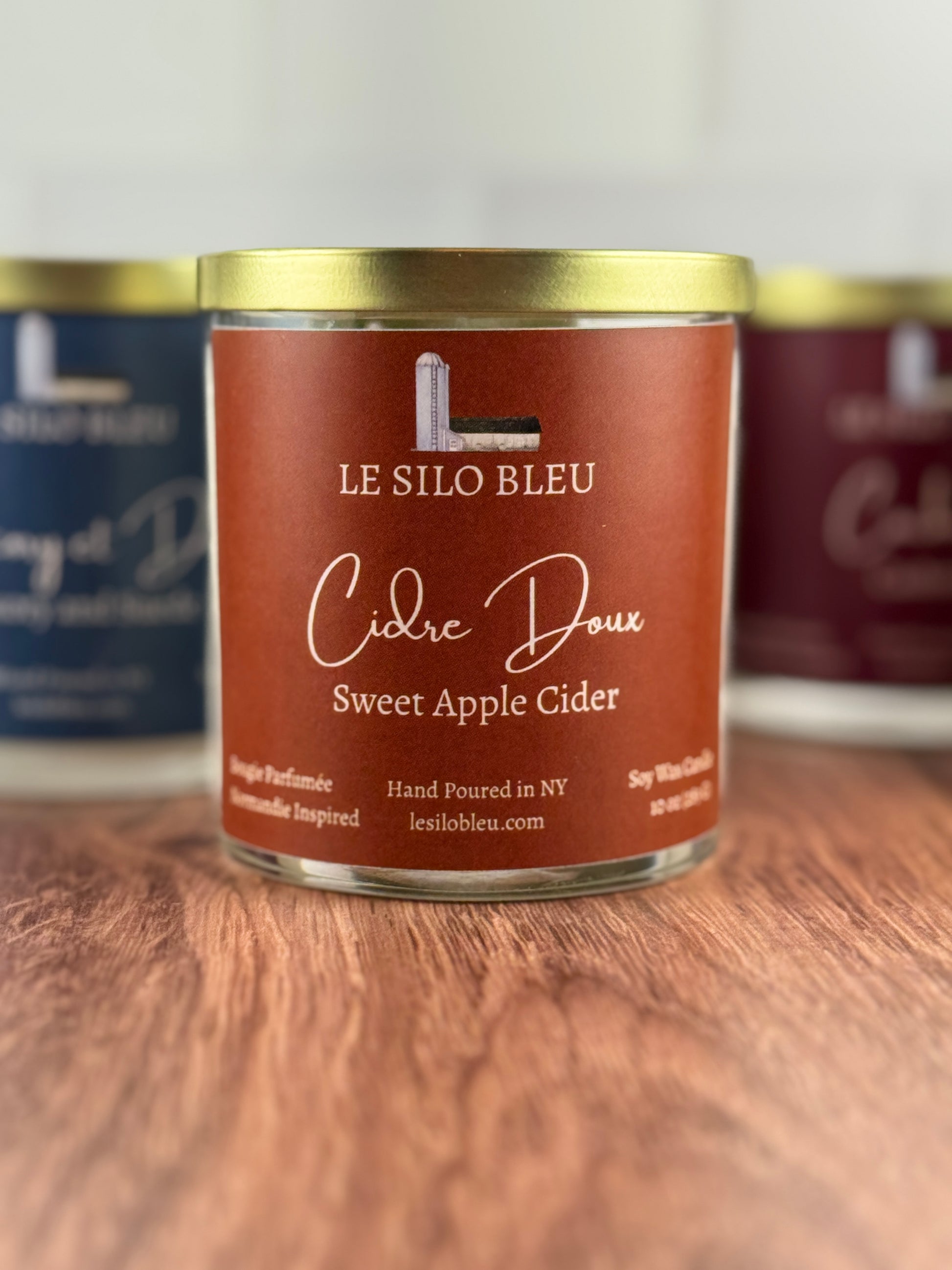 Candle in a glass jar with a gold lid labeled 'Le Silo Bleu Cidre Doux Sweet Apple Cider' on a wooden surface.