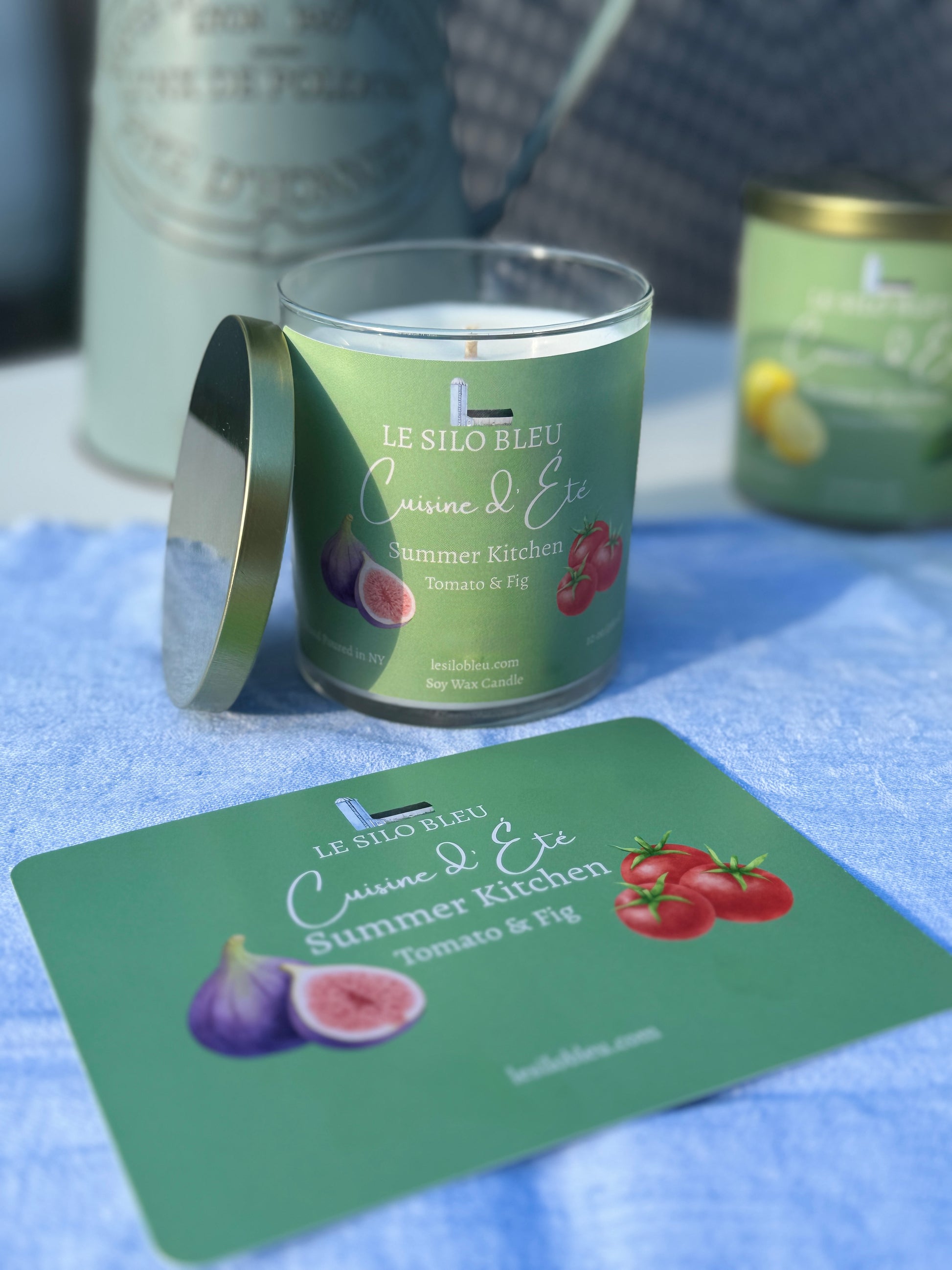 Summer Kitchen Tomato and Fig Candle with a green label on a blue surface, surrounded by similar candles and a pitcher of flowers in the background. 