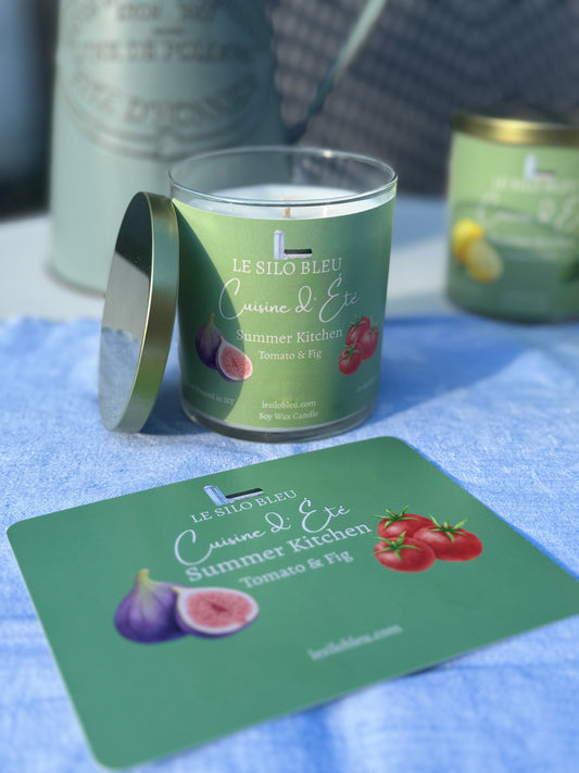 Summer Kitchen Tomato and Fig Candle with a green label on a blue surface, surrounded by similar candles and a pitcher of flowers in the background. 