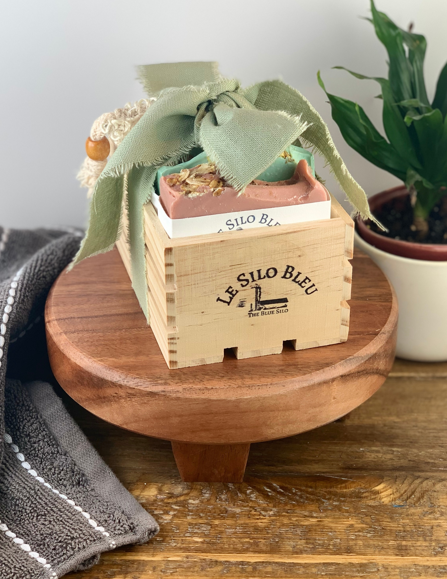 On a brown woodtopped table sits a wood stand with a small natural wood crate filled with three bars of our best selling soaps and an exfoliating scrub soap bag. This bundle is tied with a bow that is green and has the wooden box stamped with our logo. There is a green plant in a white pot in the background and a grey and white stipped hand towel on the table in the foreground. There is a white background and a wooden table top. 