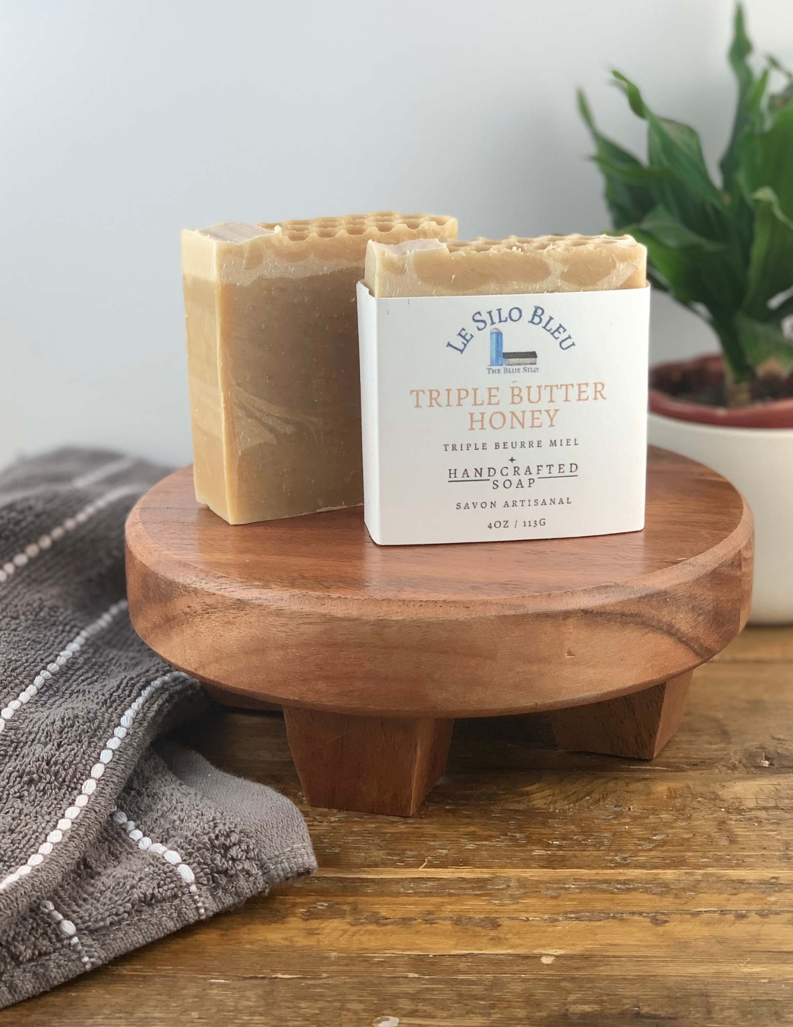 Two bars of Triple Butter Honey Soap sitting on a wooden platform with one soap wrapped in white labeling and one without. There is a green plant in a white pot in the background and a grey and white striped towel in the foreground. There is a wooden table and white background. 