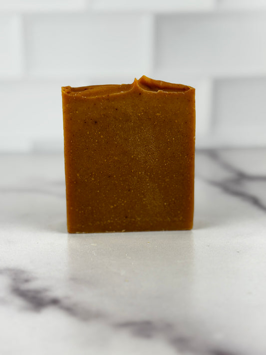 A single bar of handmade turmeric soap in a deep warm color, standing upright on a white marble surface with a white subway tile background.
