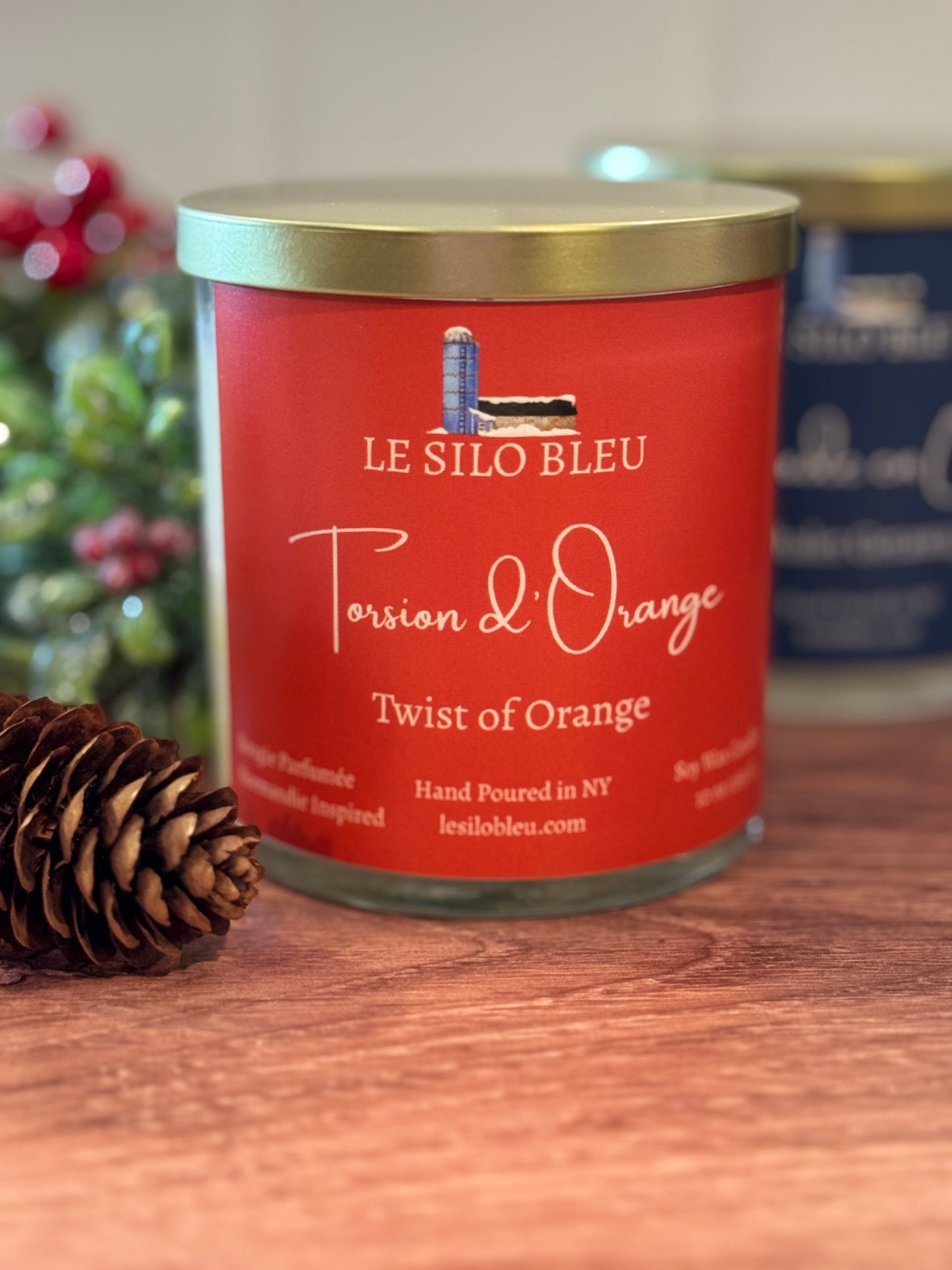 Orange labeled candle jar with gold lid labeled 'Le Silo Bleu Twist of Orange' on a wooden surface.