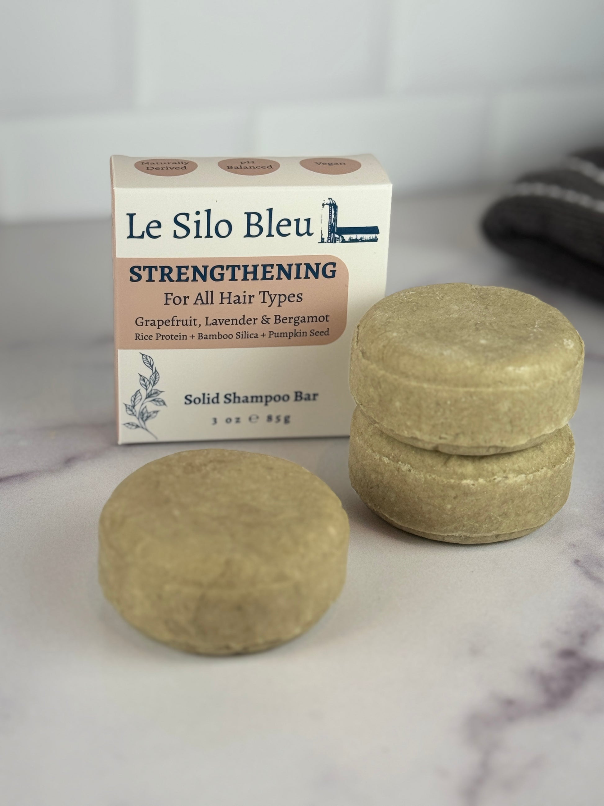 Two Strengthening solid shampoo bars stacked and one alone with their packaging on a marble surface.