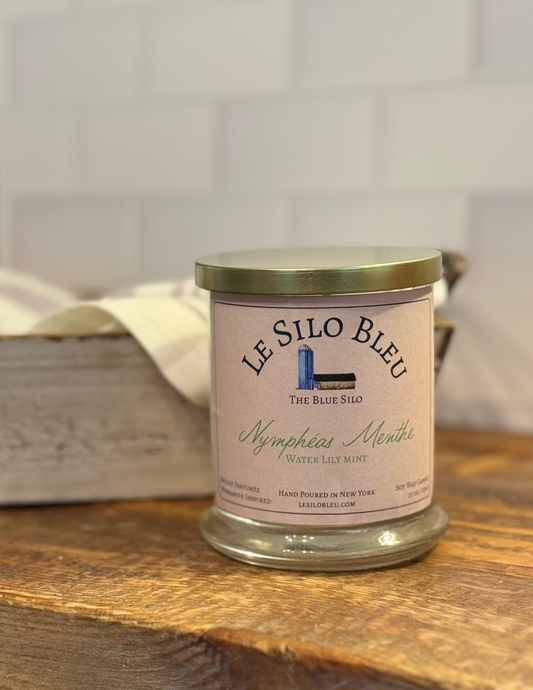 Nympheas Menthe, a fresh floral and minty scented candle with gold lid to the side. Candle is sitting on top of a wood table with an antique box and linen cloth in the background.