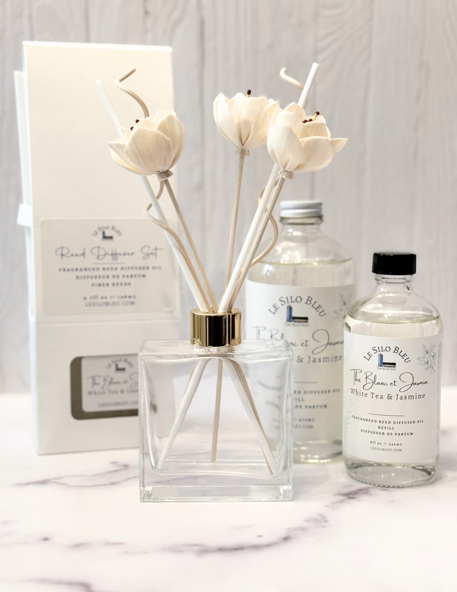 White Tea Scented Reed Diffuser set and collection showcasing the optional floral reeds along with the included white reeds and additional for purchase bottles of 8oz or 16oz fragrance refills.  The whole scent collection is sitting on a white marble table with a white wood background. 