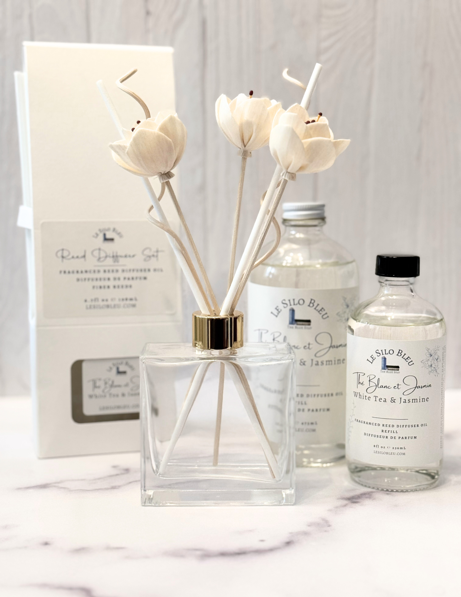 White Tea Scented Reed Diffuser set and collection showcasing the optional floral reeds along with the included white reeds and additional for purchase bottles of 8oz or 16oz fragrance refills.  The whole scent collection is sitting on a white marble table with a white wood background. 