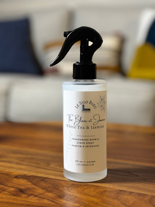 A bottle of Le Silo Bleu White Tea & Jasmine room spray, featuring a black trigger spray top and a cream-colored label, set on a wooden table with a blurred living room background.
