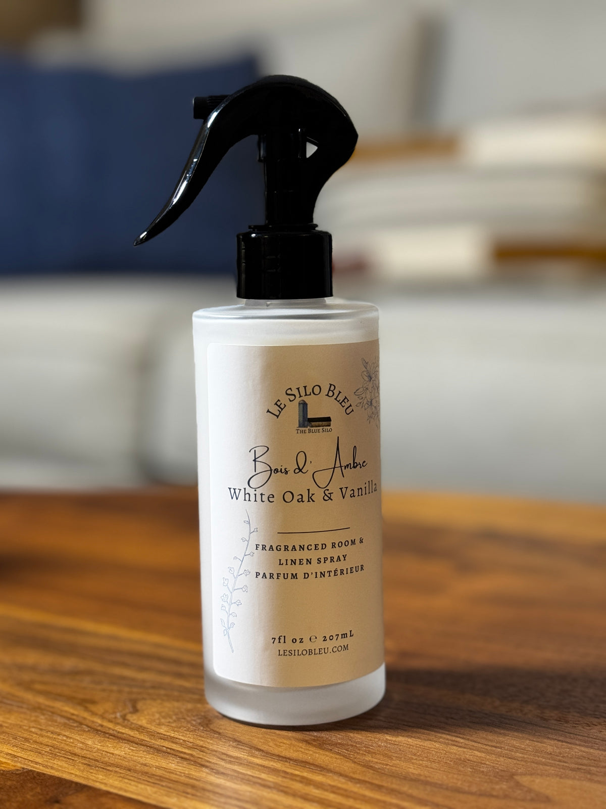 White Oak and Vanilla room and linen spray in a white frosted bottle sitting on a walnut table with a blurred living room sofa in the background. 