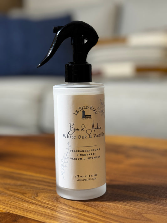 White Oak and Vanilla room and linen spray in a white frosted bottle sitting on a walnut table with a blurred living room sofa in the background. 