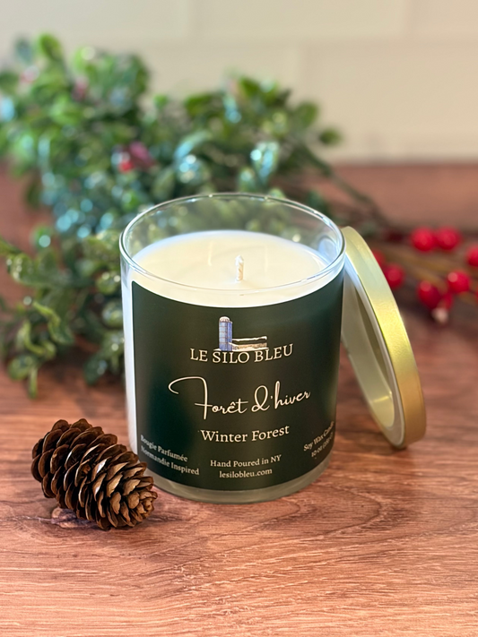 Candle in a glass jar with a dark green label on a wooden surface, surrounded by decorative elements of pinecones and greenery.