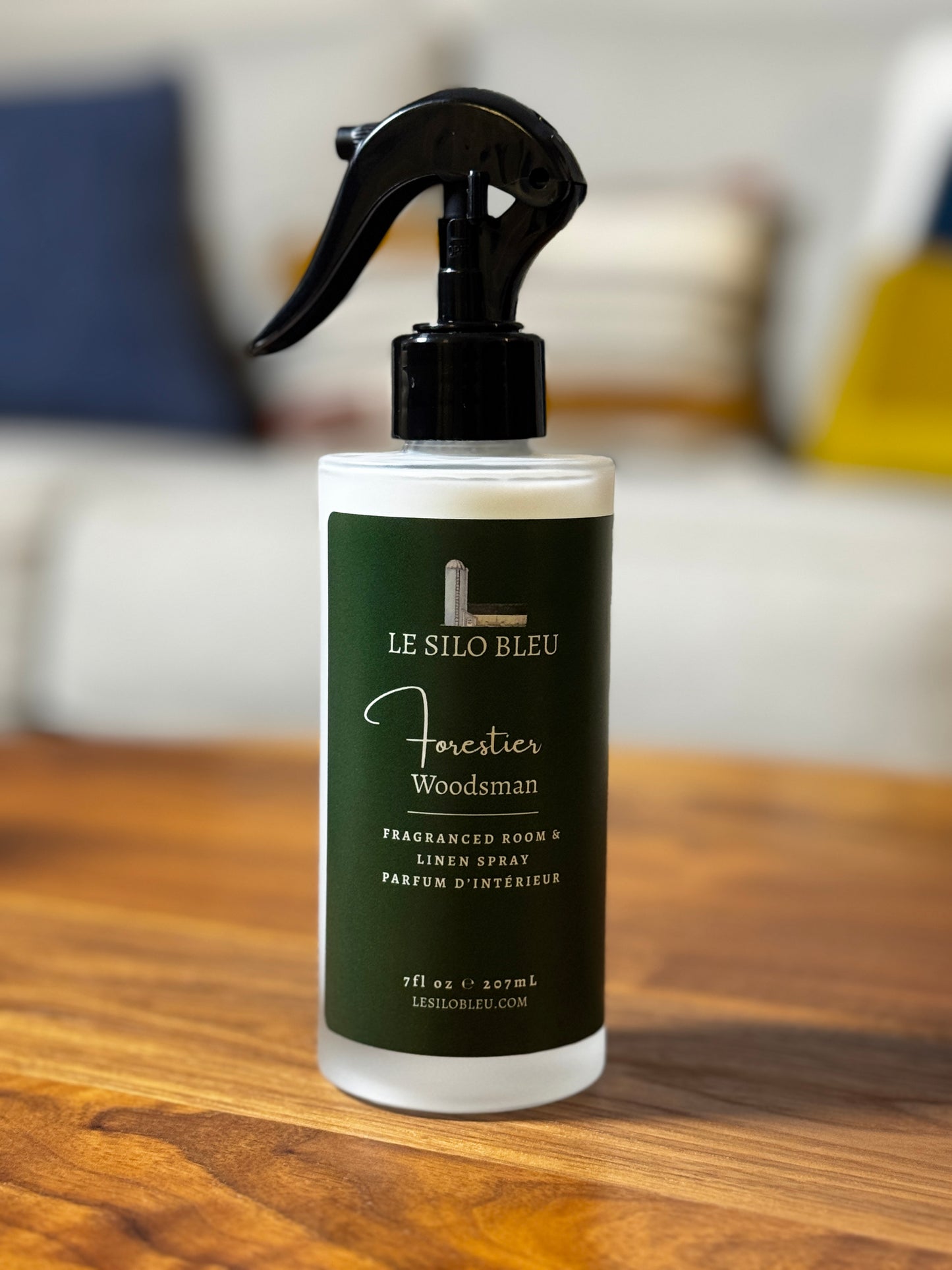 A bottle of Le Silo Bleu Forester - Woodsman Fraganced Room & Linen Spray with a black trigger sprayer and dark green label, sitting on a wooden table with a blurred living room background.