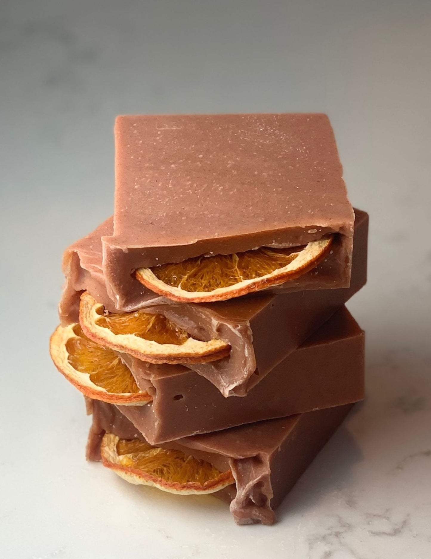 Stack of four dark orange soaps. Details the dried orange slice on the top. All soaps are sitting on a marble counter with a white backsplash. 