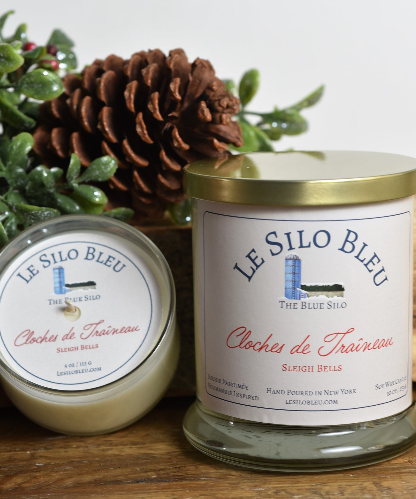 Sleigh Bells Candle Duo- Large and Small Candles sitting on a wooden table with a pine cone and green holly berry leaves and berries in the background.