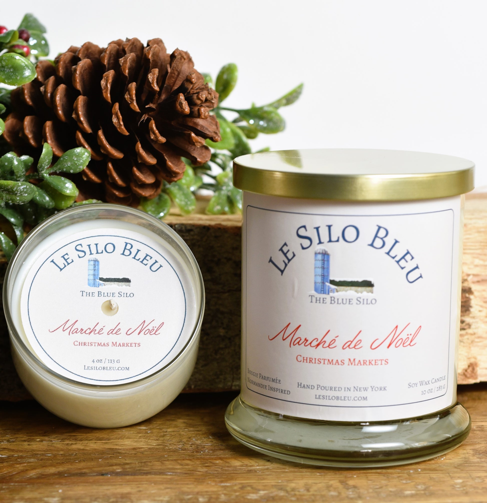 Christmas Markets Candle Duo-Large and Small Candles sitting on a wooden table with a pine cone and green holly berry leaves and berries in the background.