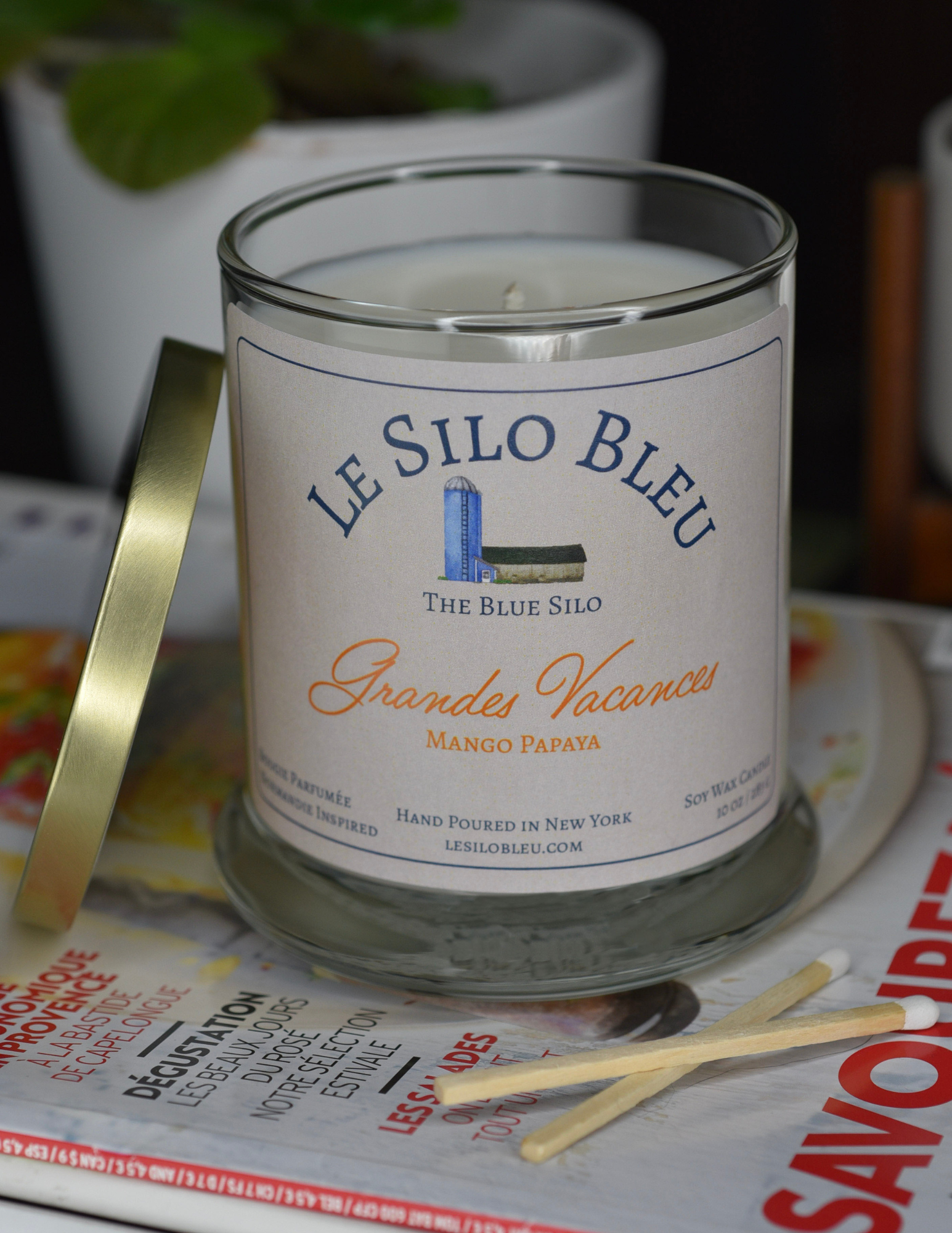 Grandes Vacanes, a mango papaya  scented candle with gold lid to the side. Candle is sitting on top of a French magazine with a plant in a white pot in the background.