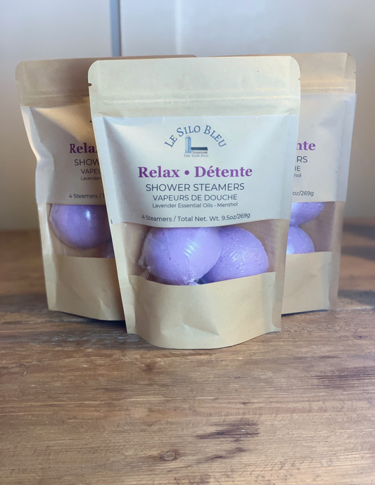 Three packages of Relax Detente Shower Steamers sit on a wooden table with a white bead board background. Two purple steamers are visible with the package windows.